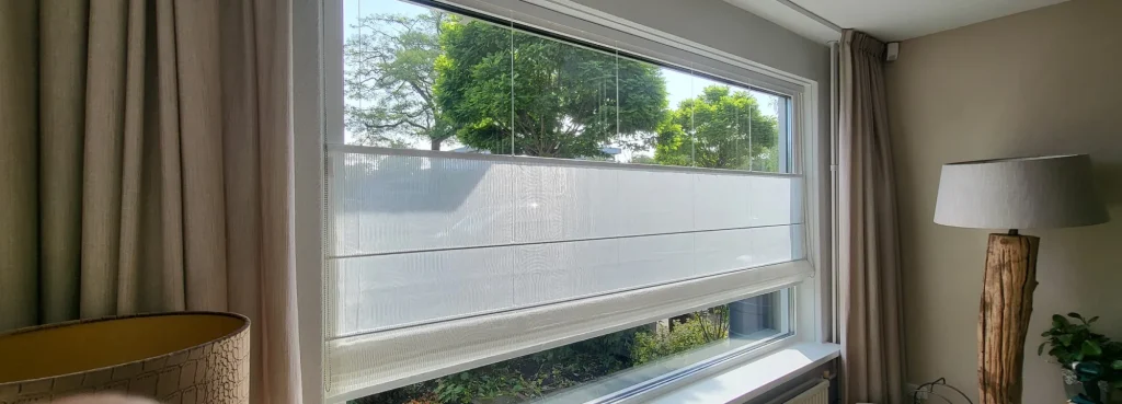 Een groot raam met transparante vouwgordijn top down bottom up, beige gordijnen aan de zijkanten, een zichtbare boom buiten en een houten lamp in de hoek van de kamer.