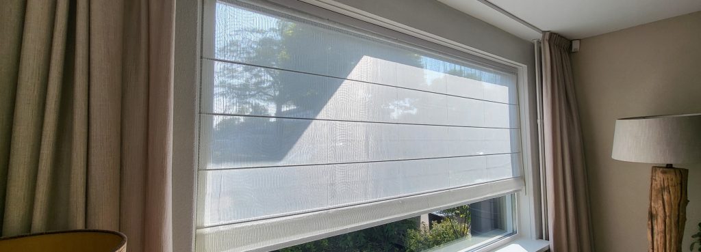 Een groot raam met doorschijnende witte vouwgordijnen top down bottom up, gedeeltelijk neergelaten, laat natuurlijk licht binnen. Beige gordijnen omlijsten het raam en rechts staat een staande lamp.