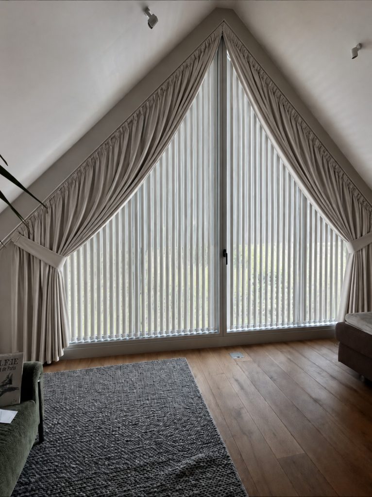 Een driehoekig raam met trapezium gordijnen op maat en gedrapeerde gordijnen in een kamer met houten vloer, een grijs vloerkleed, een groene stoel en een bruine bank.